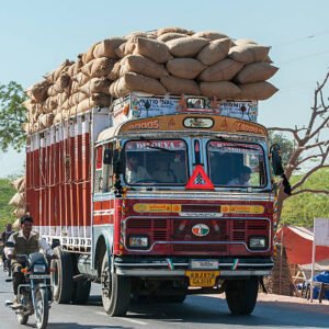 Nagaur, India - February 10, 2011: Overloaded dump truck filled with jute bags on a rural road. Driver and helper in truck. Other traffic are motorcyclists and a pedestrian.